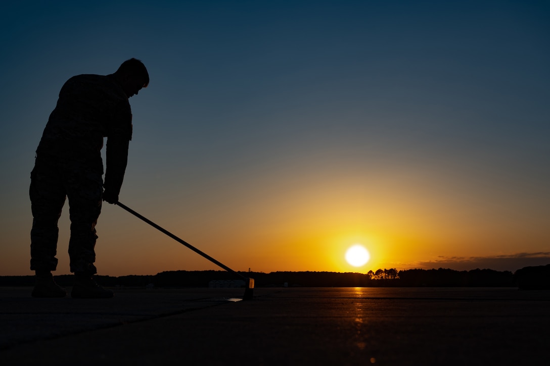 An airmen brushes concrete under a sunlit sky as shown by silhouette.