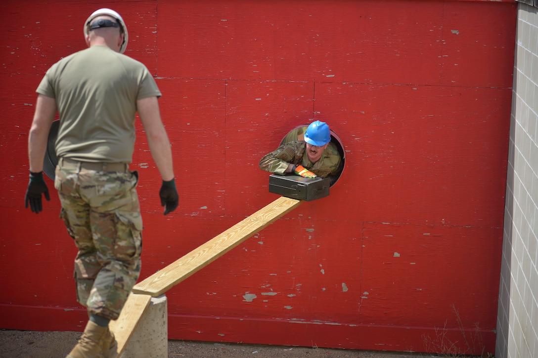 An airman climbs from a hole in a wall onto a plank while a fellow airman watches.