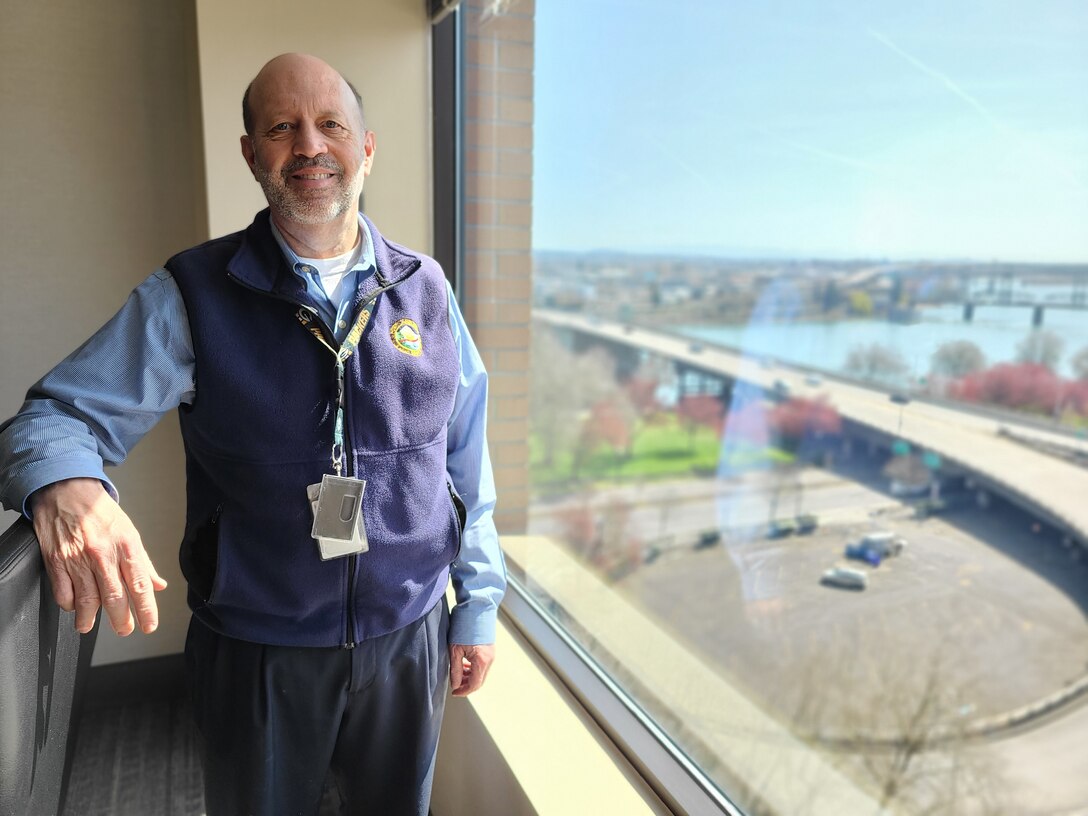 A man in a blue shirt stands at his office window, which overlooks a river.