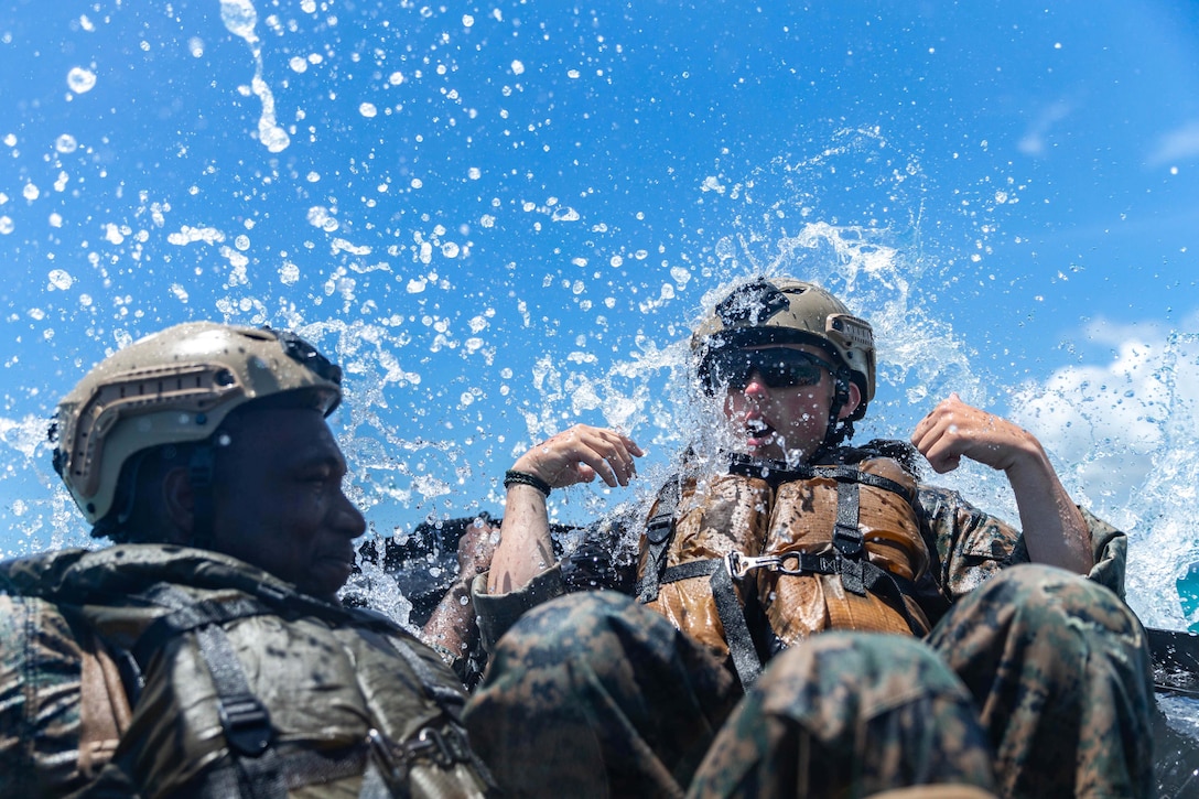Two Marines sit in a raft as water splashes around them.