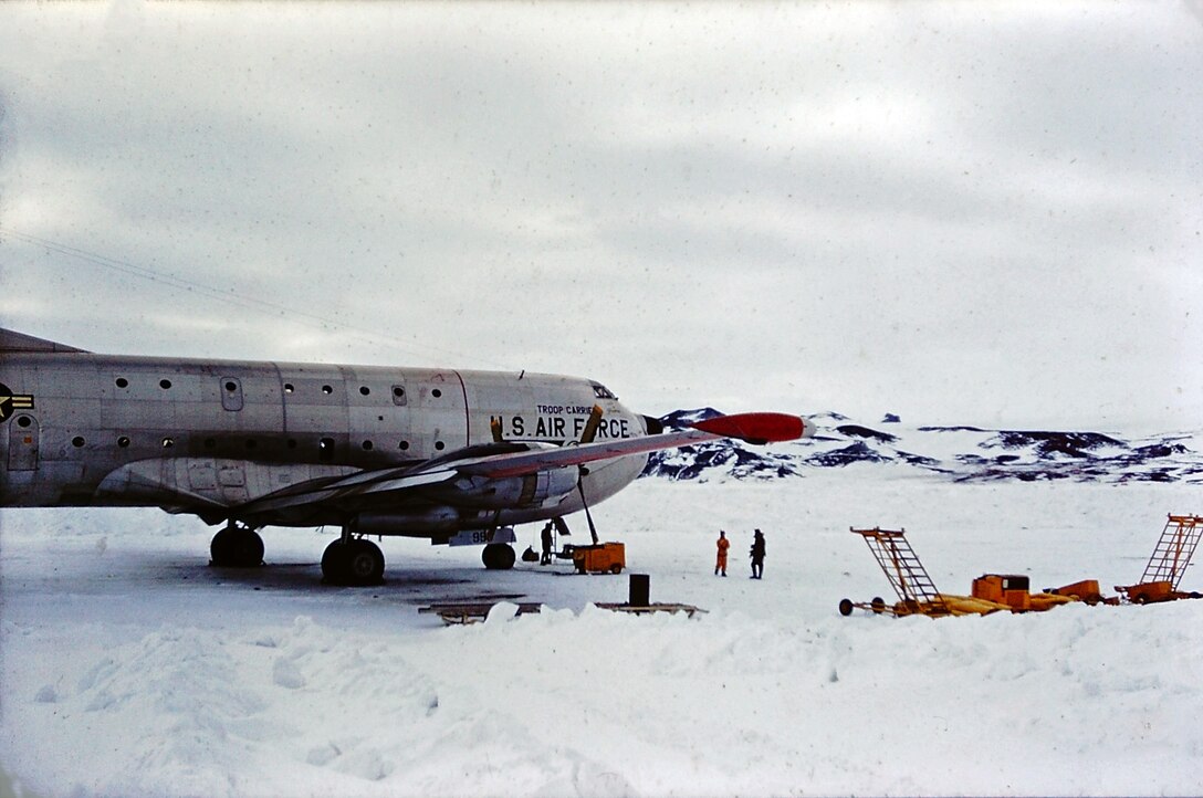A cargo plane sits on a snowed-over runway with equipment around it. Small mountains rise in the background.