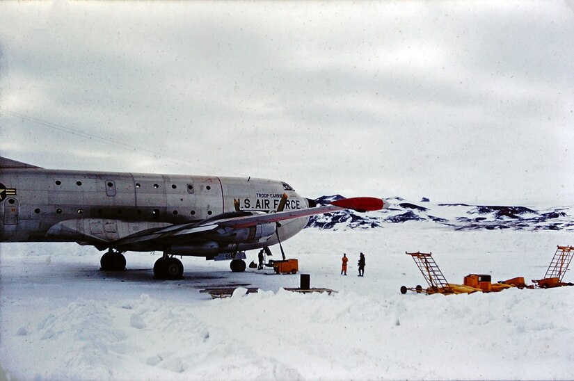 A cargo plane sits on a snowed-over runway with equipment around it. Small mountains rise in the background.