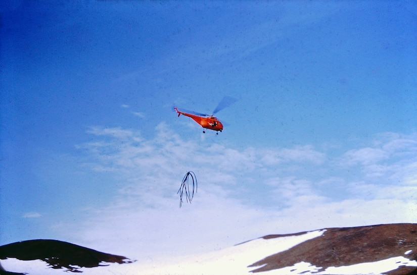 A helicopter hauls several long dangly items from its cable over a mountain.
