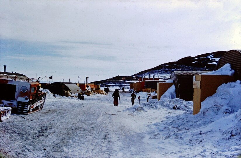 Two people walk a small snow-covered road surrounded by small buildings and a mountain.