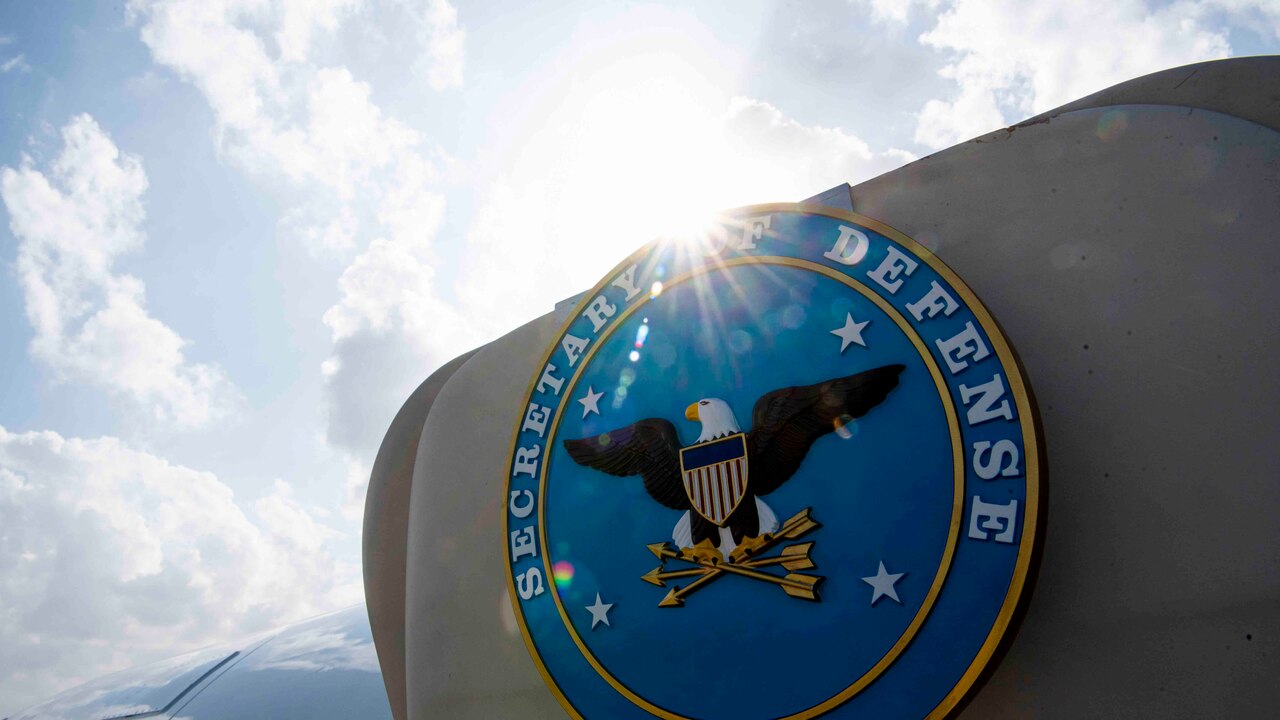 A light blue seal with an eagle and the words "SECRETARY OF DEFENSE" adorns an open aircraft door.