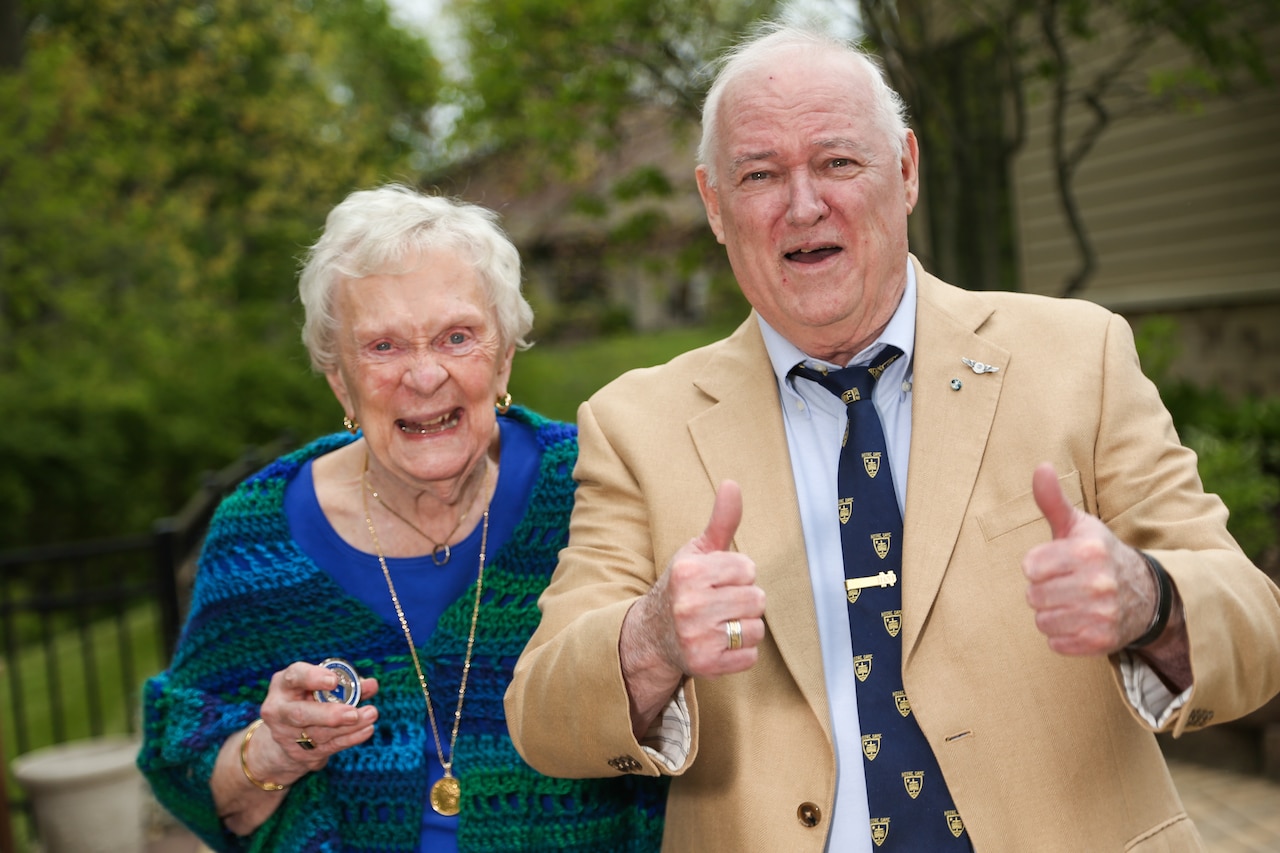 An older woman smiles while holding a coin; an older man beside her holds two thumbs up.