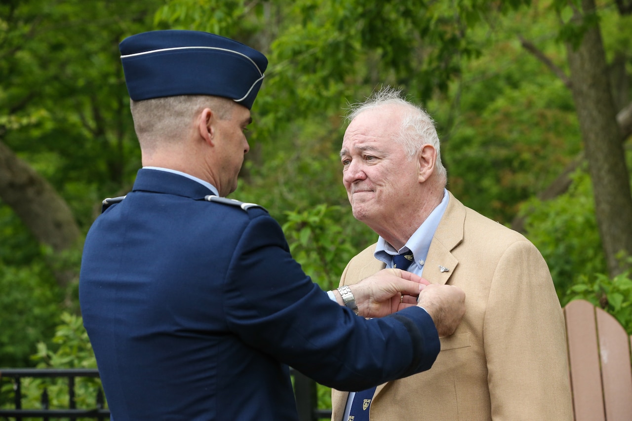 A man in a suit jacket receives a medal chest pin from a man in military uniform.