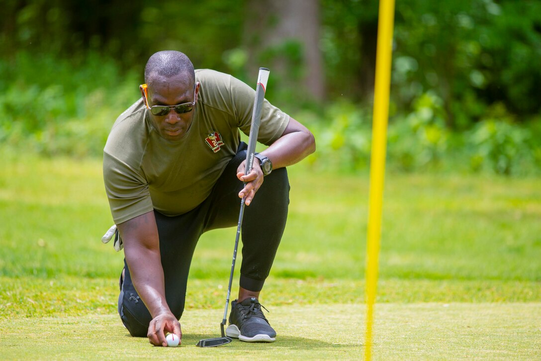 A Marine kneels on the ground while holding a golf club and a golf ball.