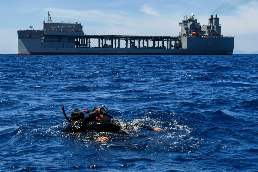 Two sailors wearing dive gear move through waters as a ship sails behind.