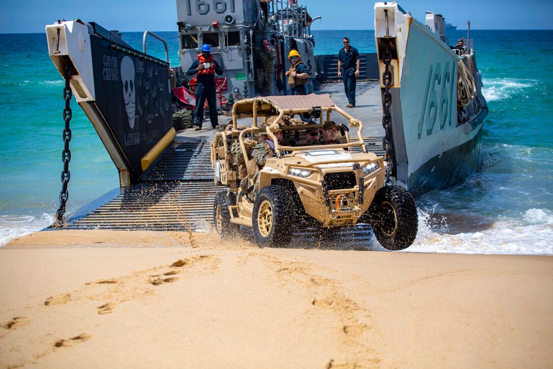 A tactical vehicle drives away from a landing craft utility on a beach.