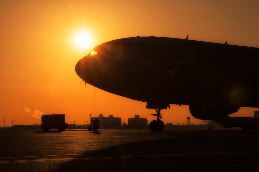 A military aircraft sits in silhouette against an orange sky and setting sun.