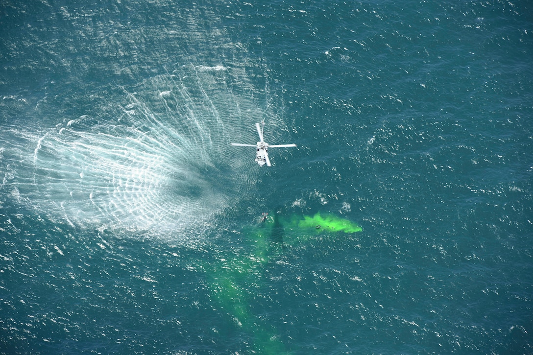 A helicopter flies over a body of water while carrying a Marine on a rope over a cloud of green smoke.