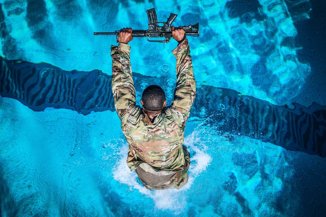 A soldier jumps into a pool while holding up a rifle.