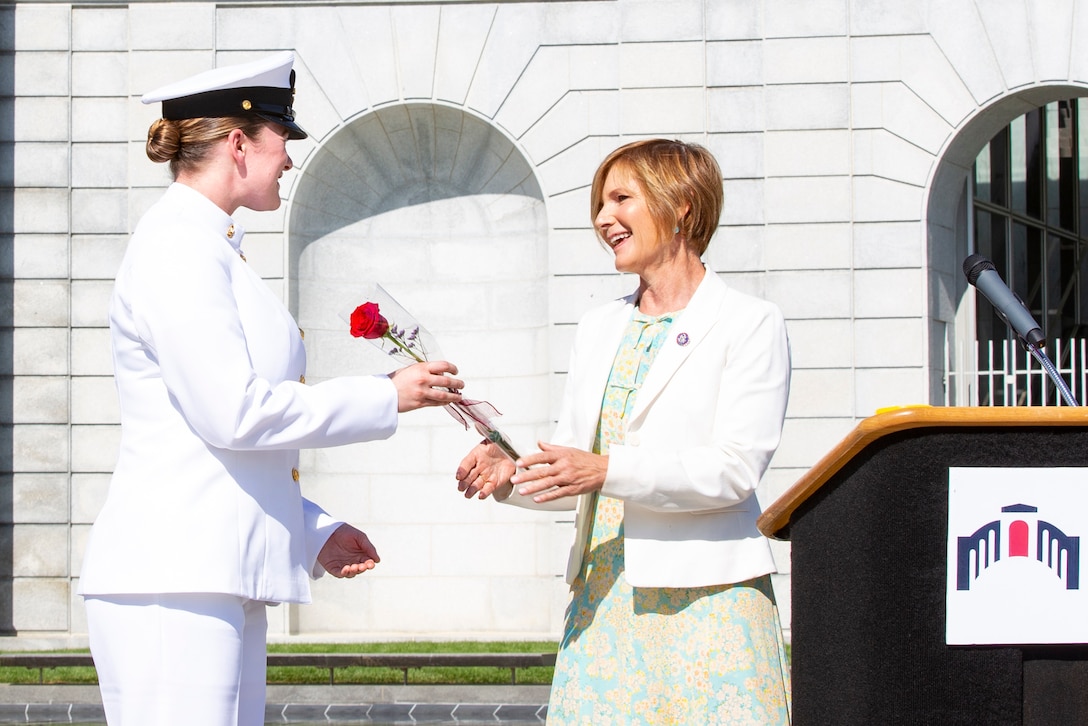 A sailor accepts a rose from a person next to a podium.