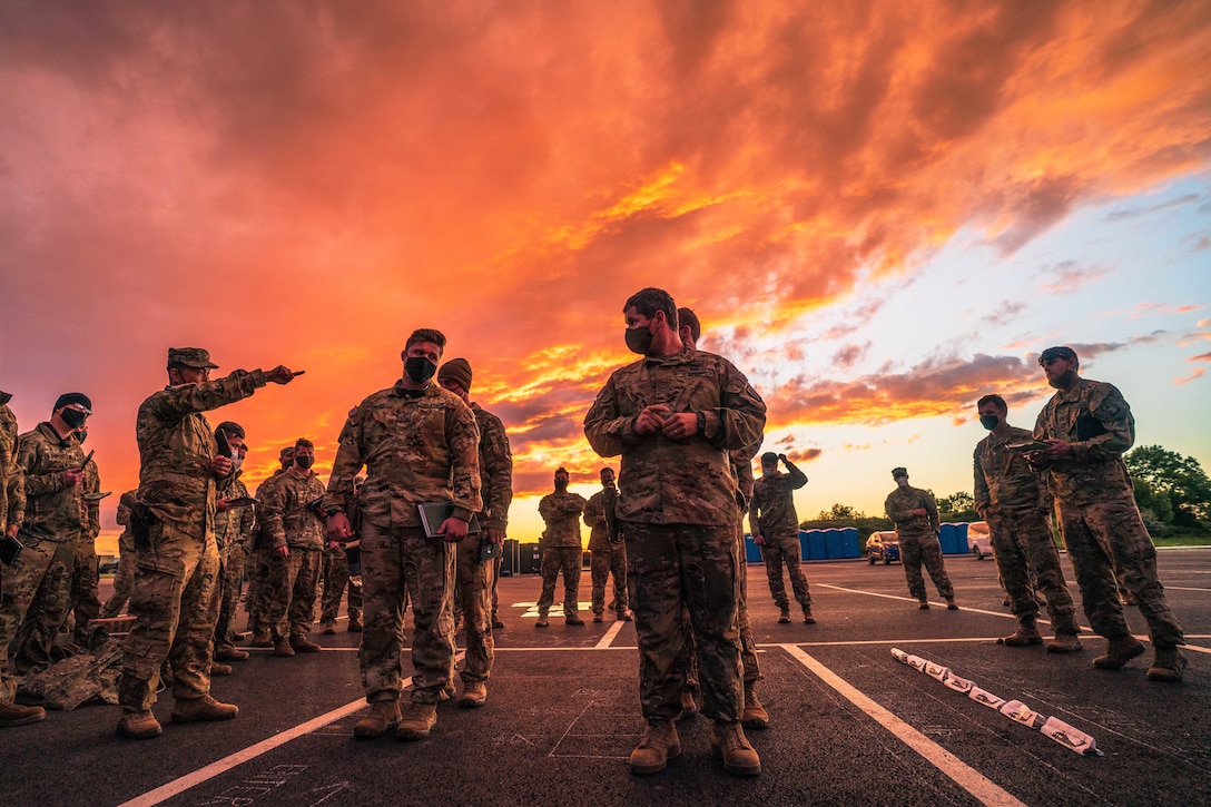 A group of soldiers gather in an open airfield under an orange-bluish sky.