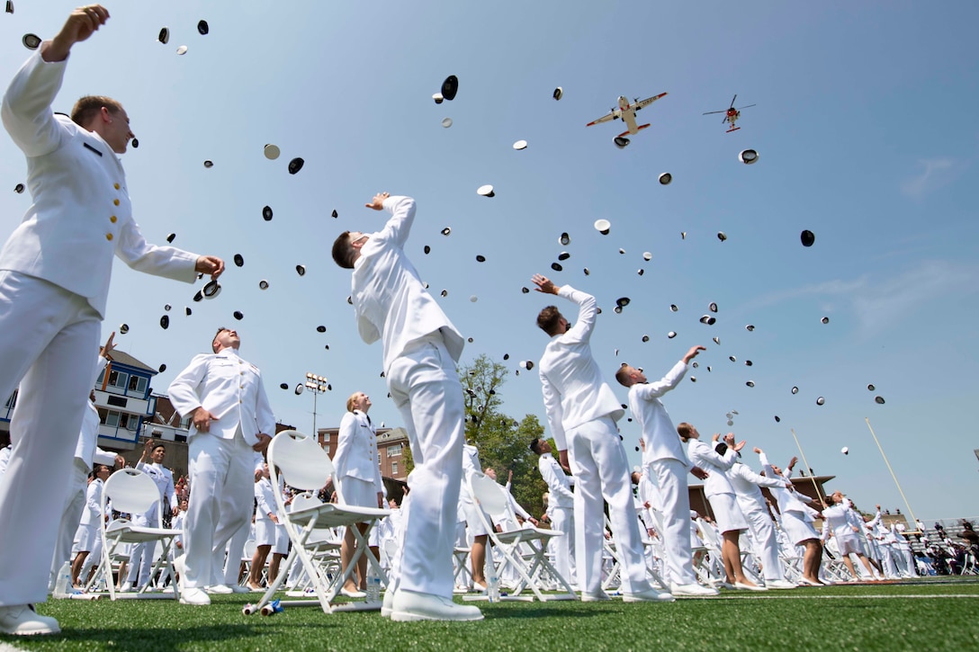 A large group of people dressed in white toss hats into the air as aircraft fly overhead.