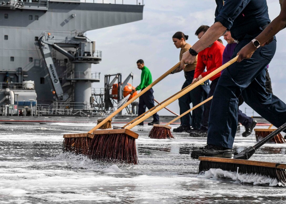 Sailors wash the deck of a ship with brooms.