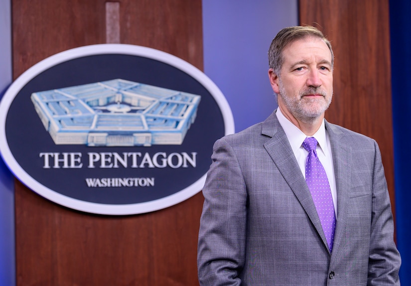 A man in a suit stands in front of a sign reading "The Pentagon - Washington."