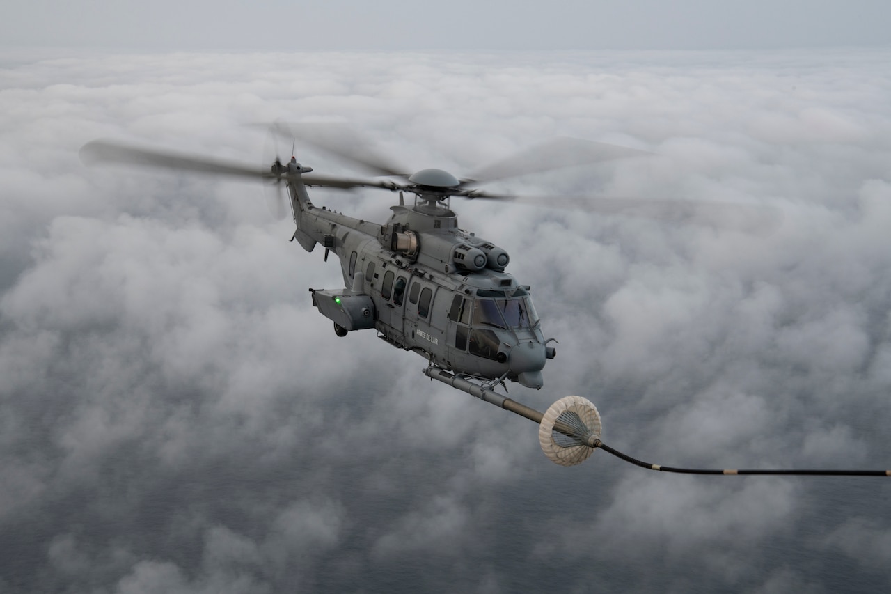 A helicopter with an attached refueling line, hovers above a body of water and clouds.