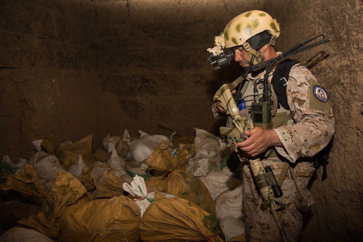 A service member with a rifle stands in a room filled with bags.
