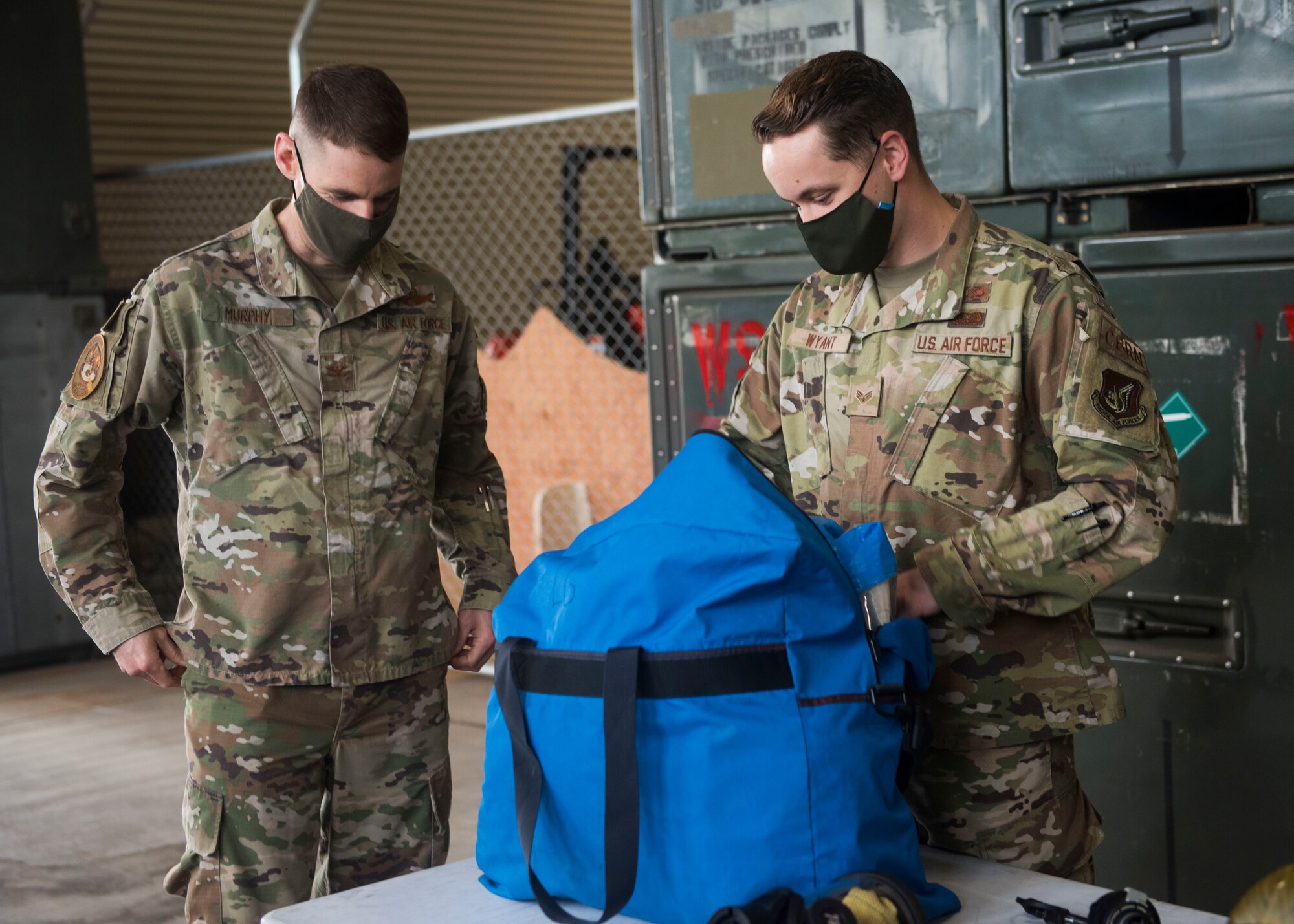 Two men in a military uniform unpack a bag containing a hazmat suit.
