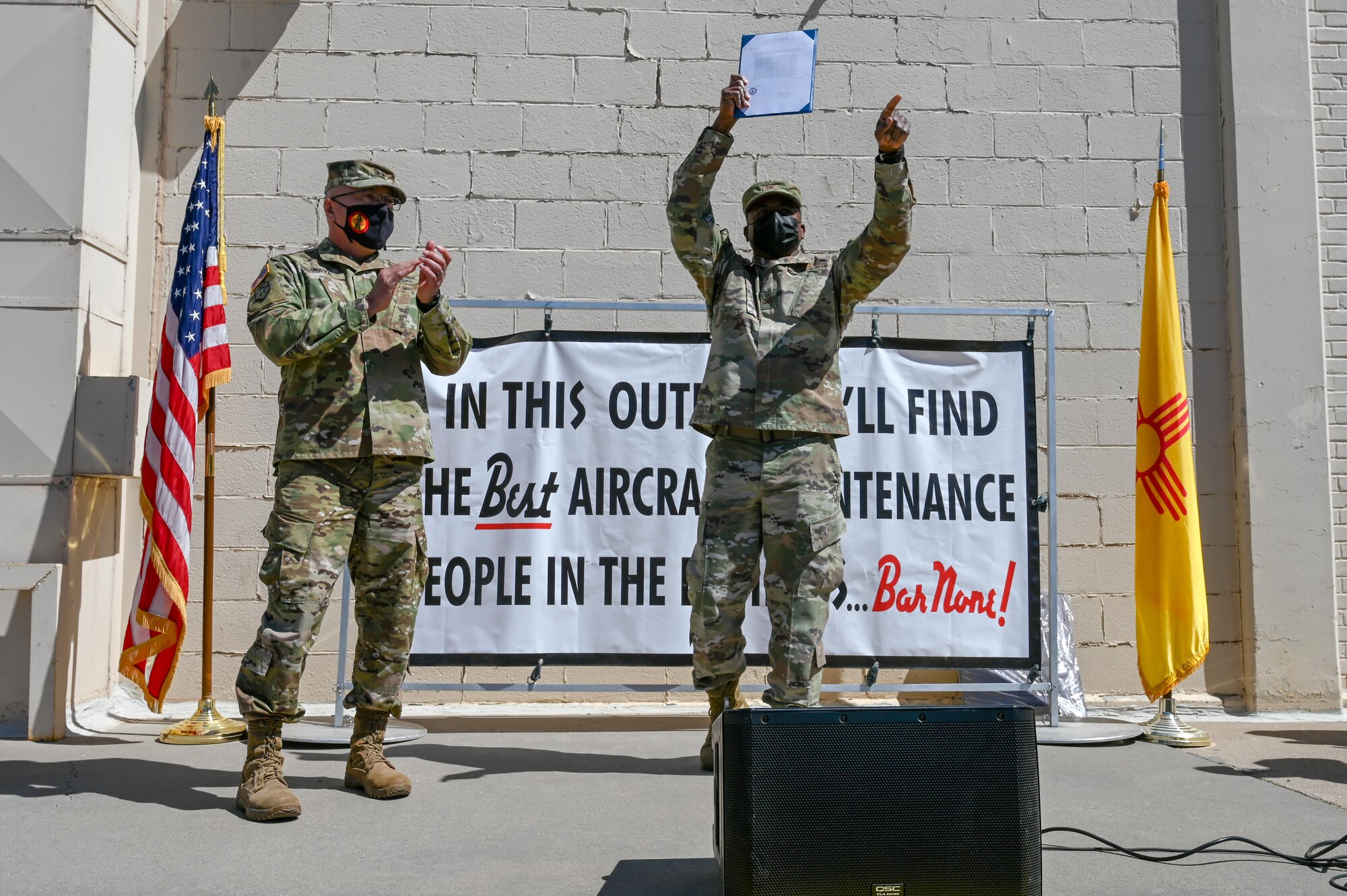 Gen. Nava and Col. Clark stand on a stage celebrating getting 2019 New Mexico State Outstanding Unit award, with a U.S. flag to the left and New Mexico state flag to the right
