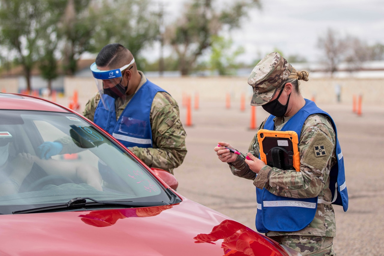 Two soldiers wearing face masks talk to a motorist in a car.