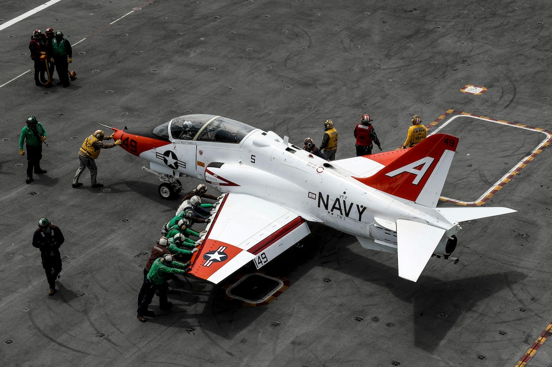 Sailors, shown from overhead, wearing uniforms of different primary colors push a plane on a flight deck.