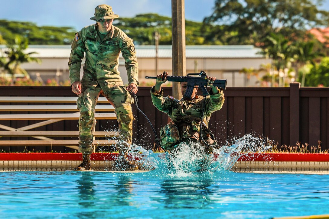A soldier jumps in a pool while carrying a weapon as a fellow soldier watches.