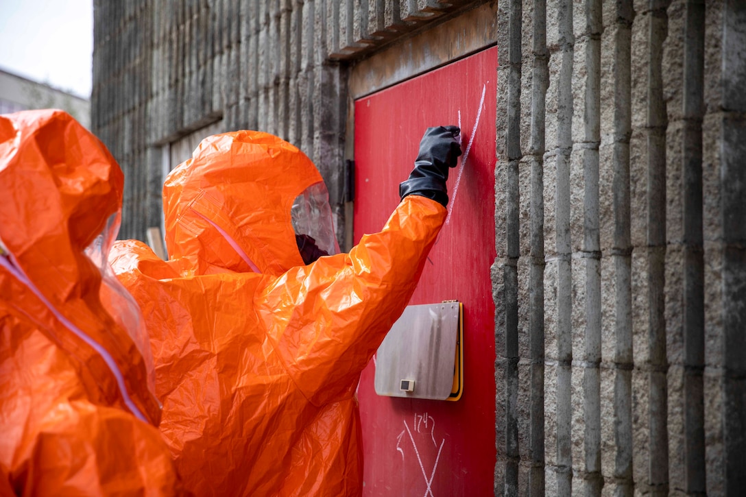 Two soldiers wearing hazmat gear stand at a door.