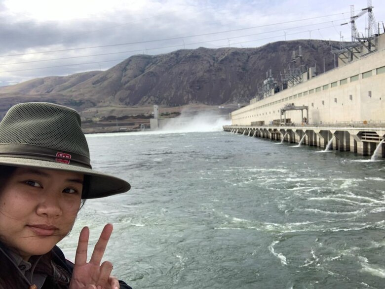 A ranger stands in front of a dam spillway, giving the peace sign.