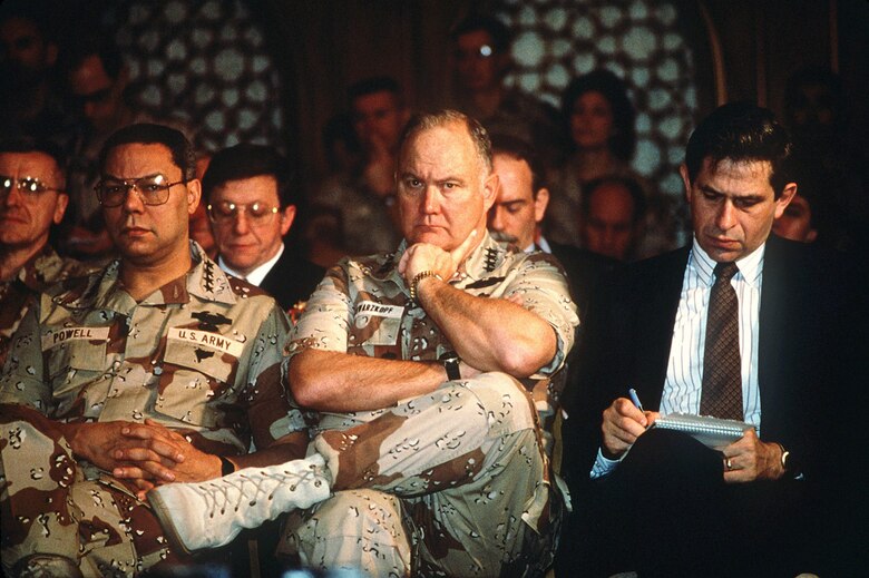 Paul Wolfowitz, under secretary of defense for policy, right, takes notes while Gen. Colin Powell, chairman, Joint Chiefs of Staff, and Gen. Norman Schwarzkopf, Jr., commander-in-chief, U.S. Central Command, listen to Secretary of Defense Dick Cheney answer questions from the media. The men are taking part in a press conference held by U.S. and Saudi Arabian officials during Operation Desert Storm.