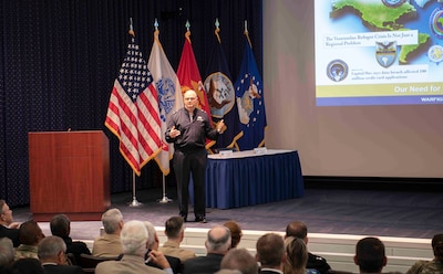 Man stands on a stage addressing an audience. Slides on the wall in the background.