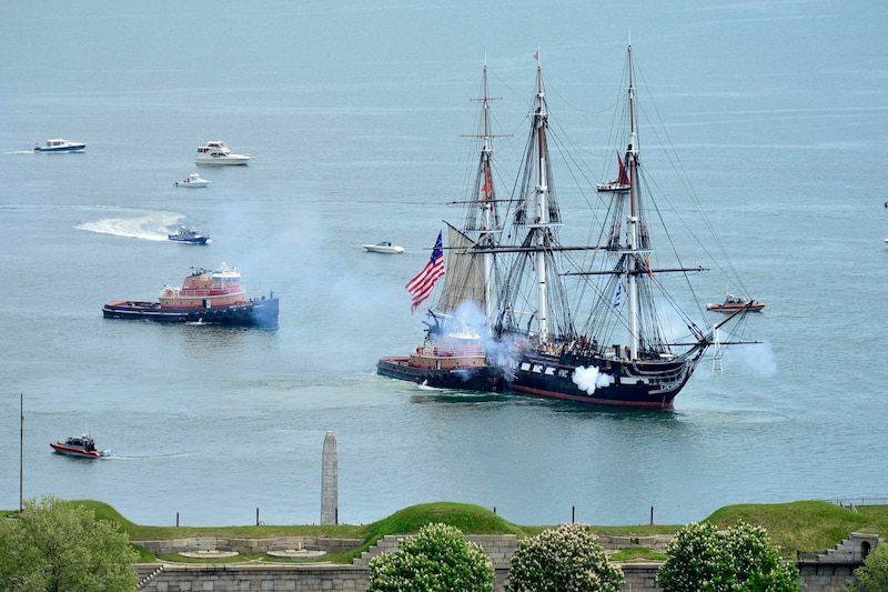 An old ship sails near two tugboats in a harbor, followed by smaller boats.