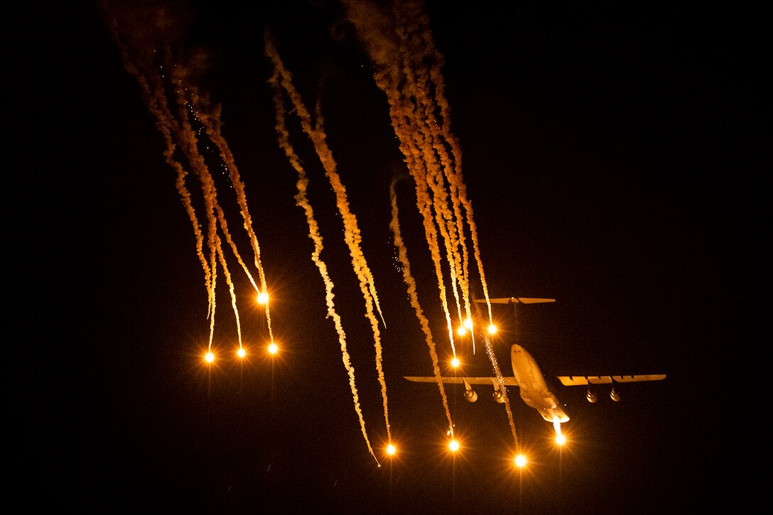 Flares illuminate a night sky as an aircraft flies nearby.