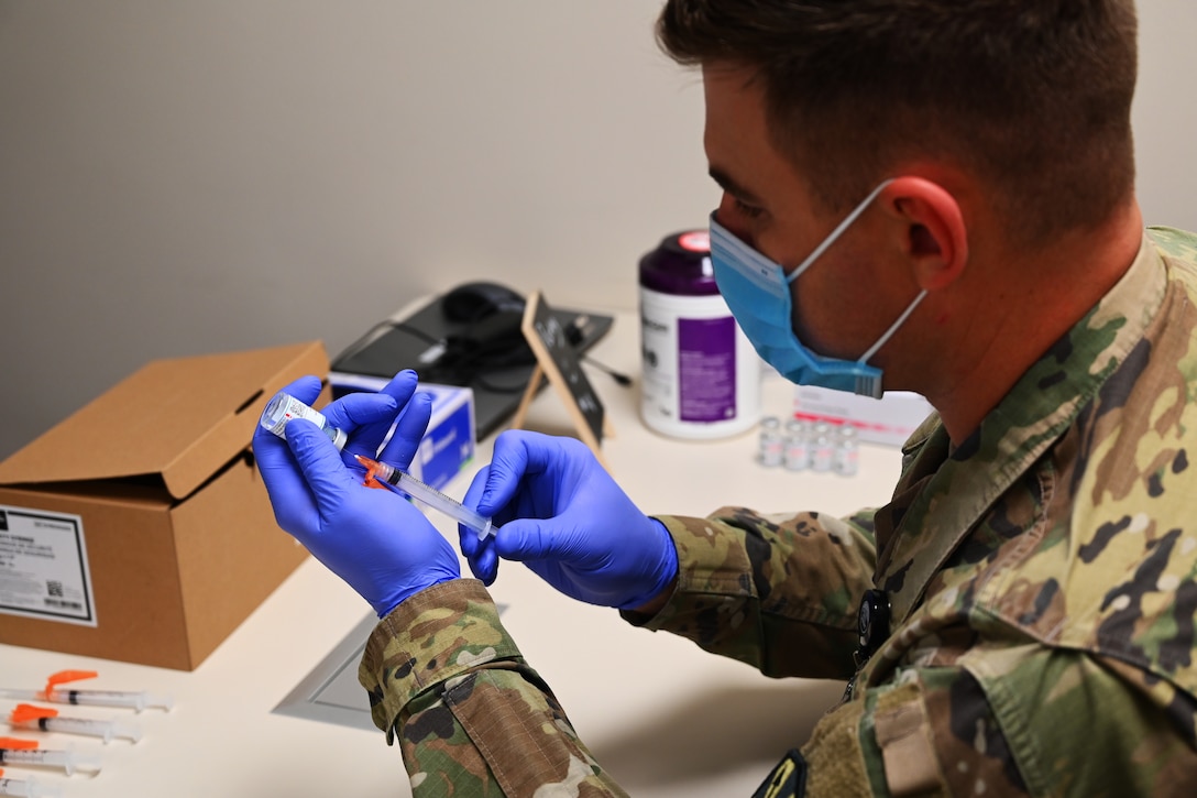 A soldier wearing a face mask and gloves holds a syringe that is being prepared for a vaccine.