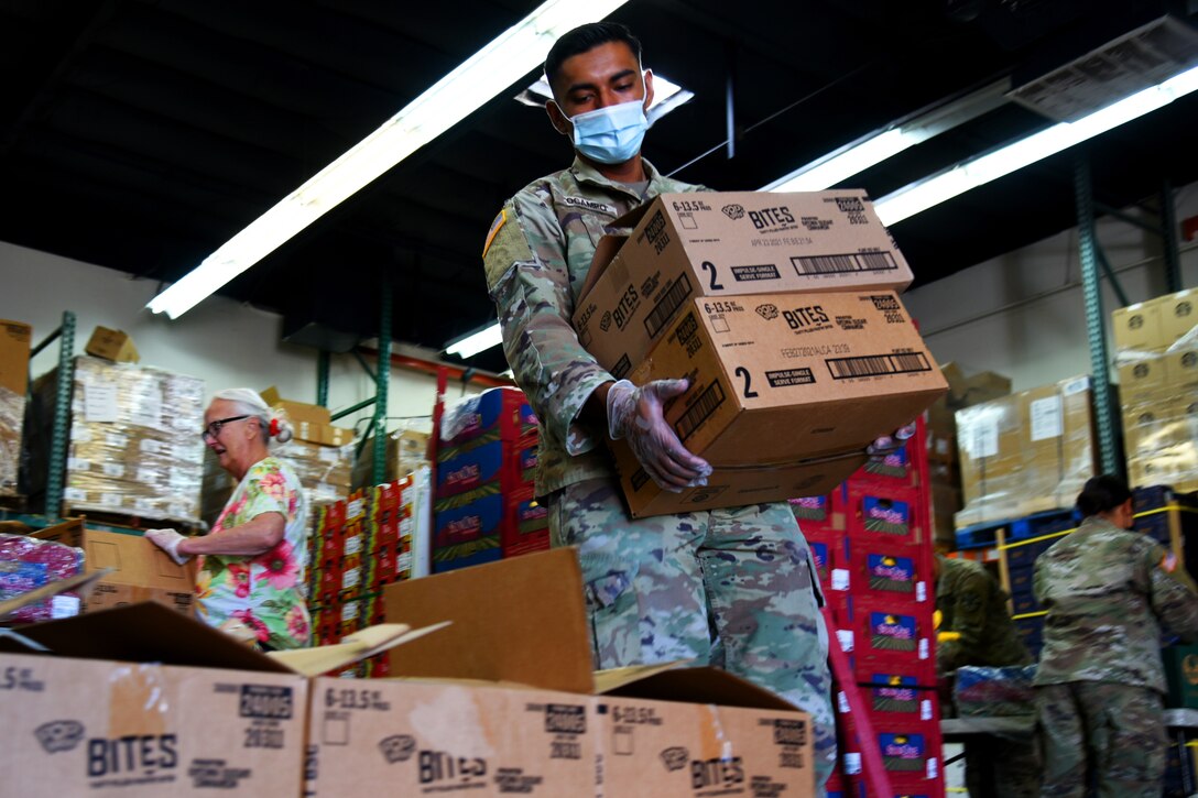 An airman wearing a face mask and gloves carries boxes of groceries that will be delivered to area residents.