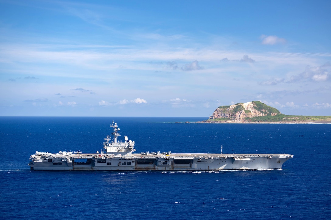 An aircraft carrier steams in the ocean with an island in the background.