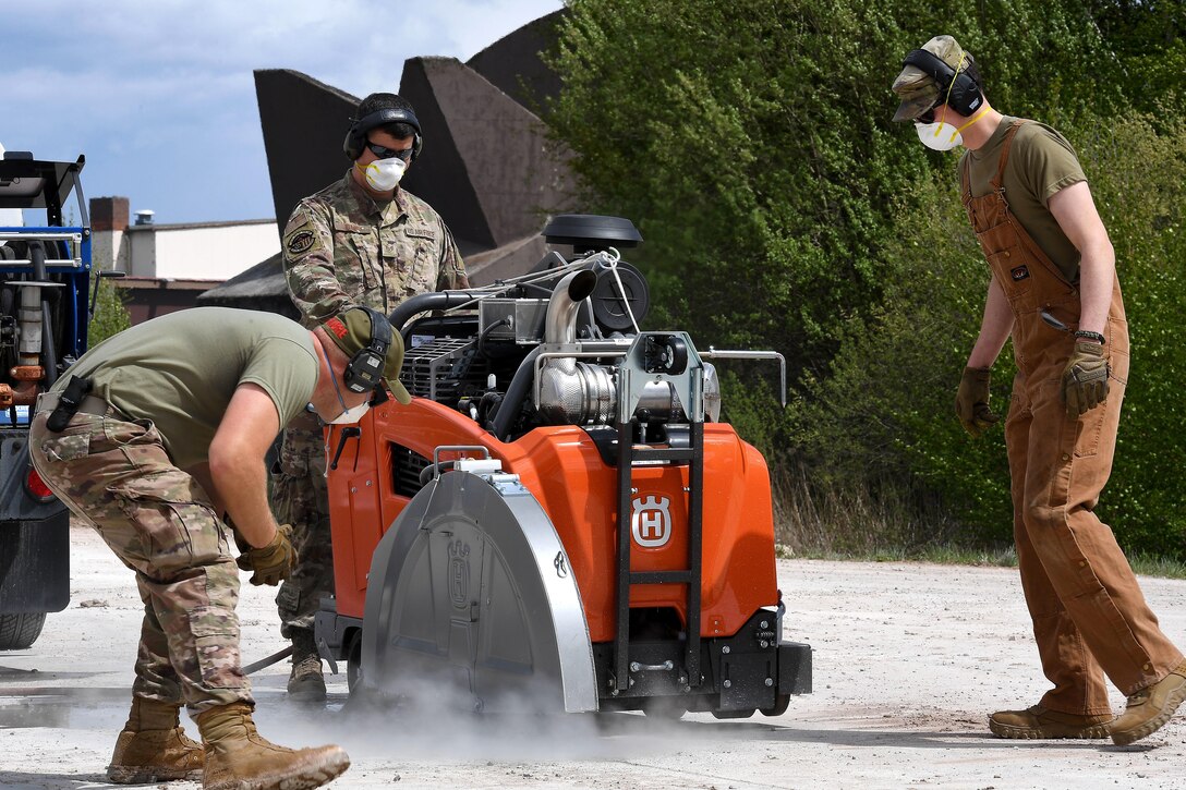 An airman rides on a construction vehicle with a wheel cutting into concrete pavement while two others monitor.