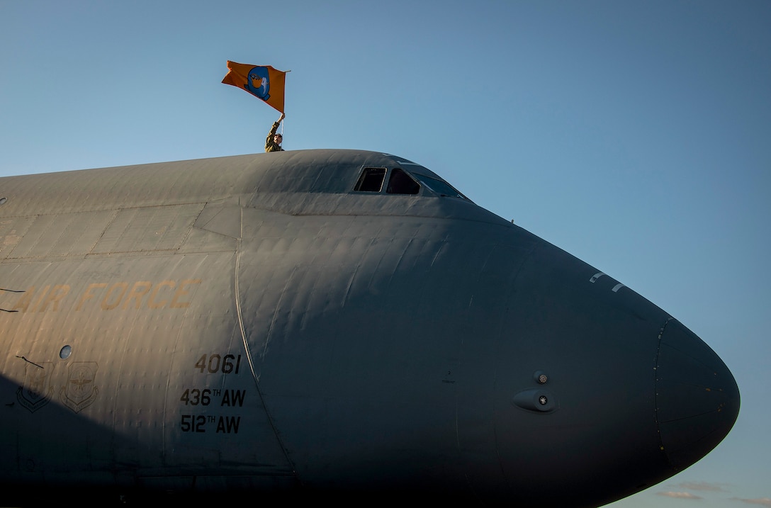 An airman waves a flag from the top of a large aircraft, against a blue sky.