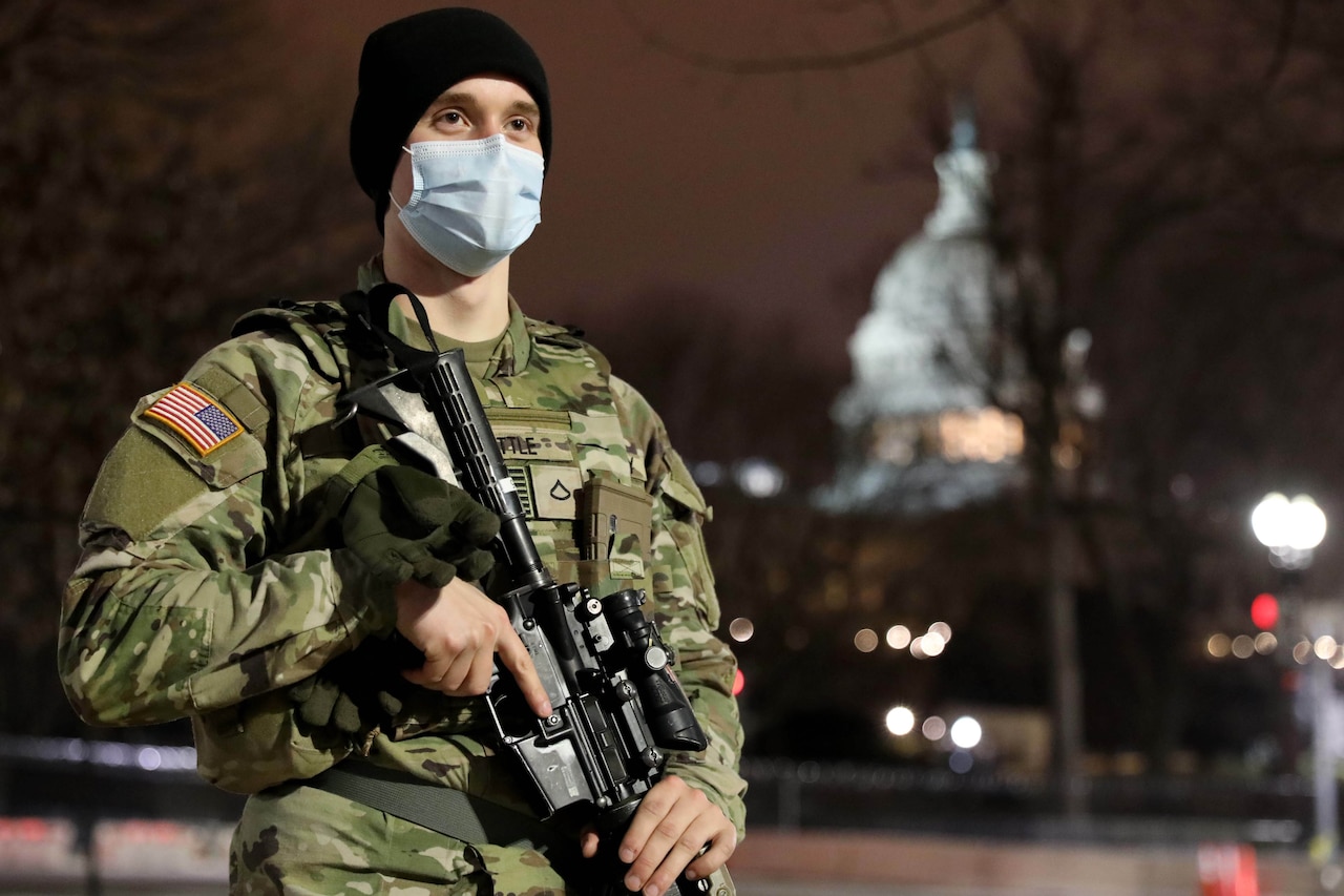 A man in a military uniform, wearing a face mask, stands with a rifle. The Capitol is in the background.