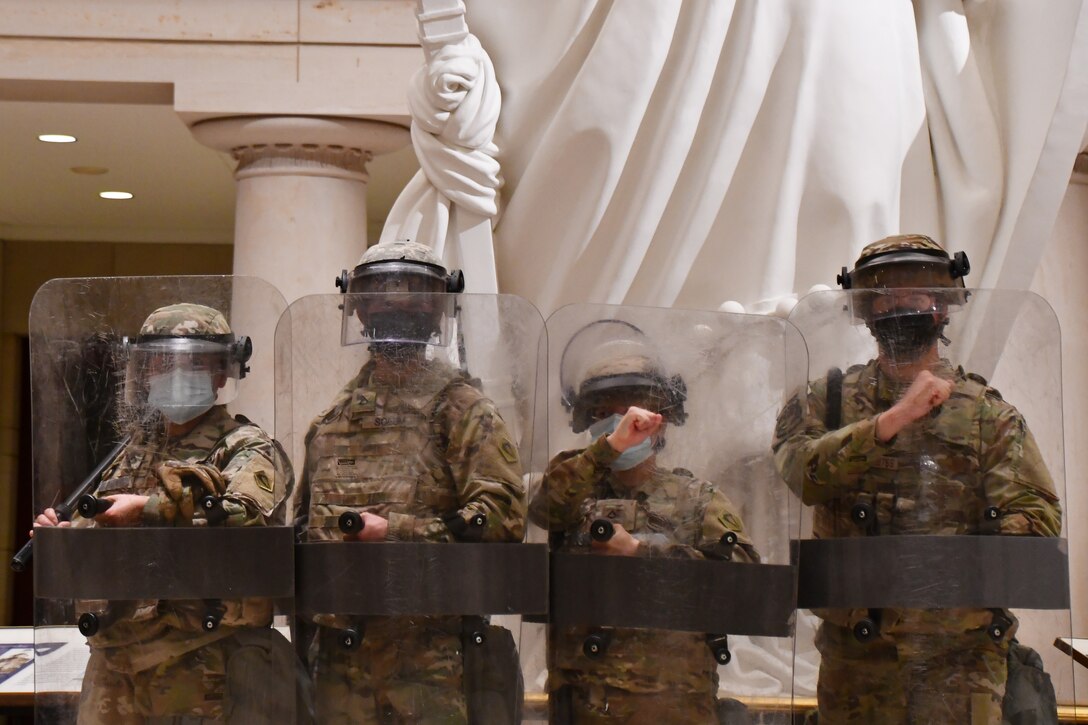 Four individuals in military uniforms stand with shields in front of a large statue.