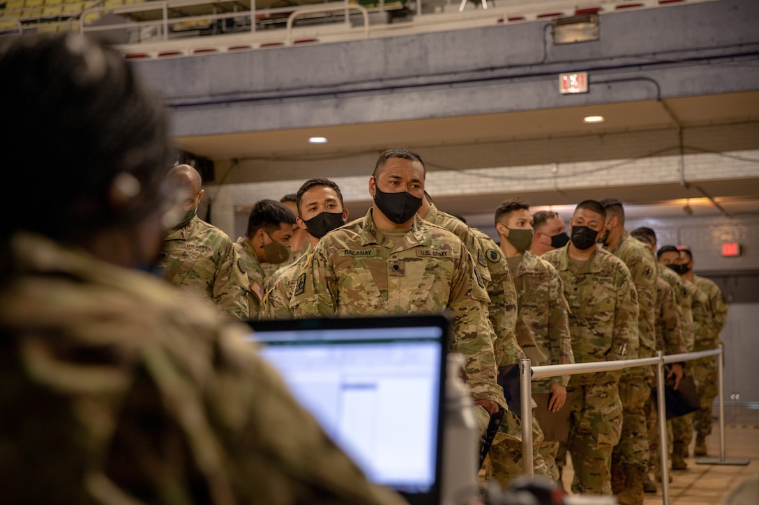 Soldiers stand in lines to approach a person with a computer.