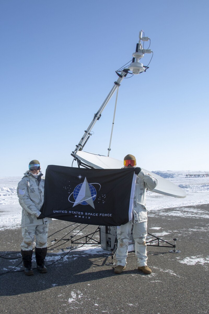 Two Space Force guardians stand in front of a satellite while holding a flag.