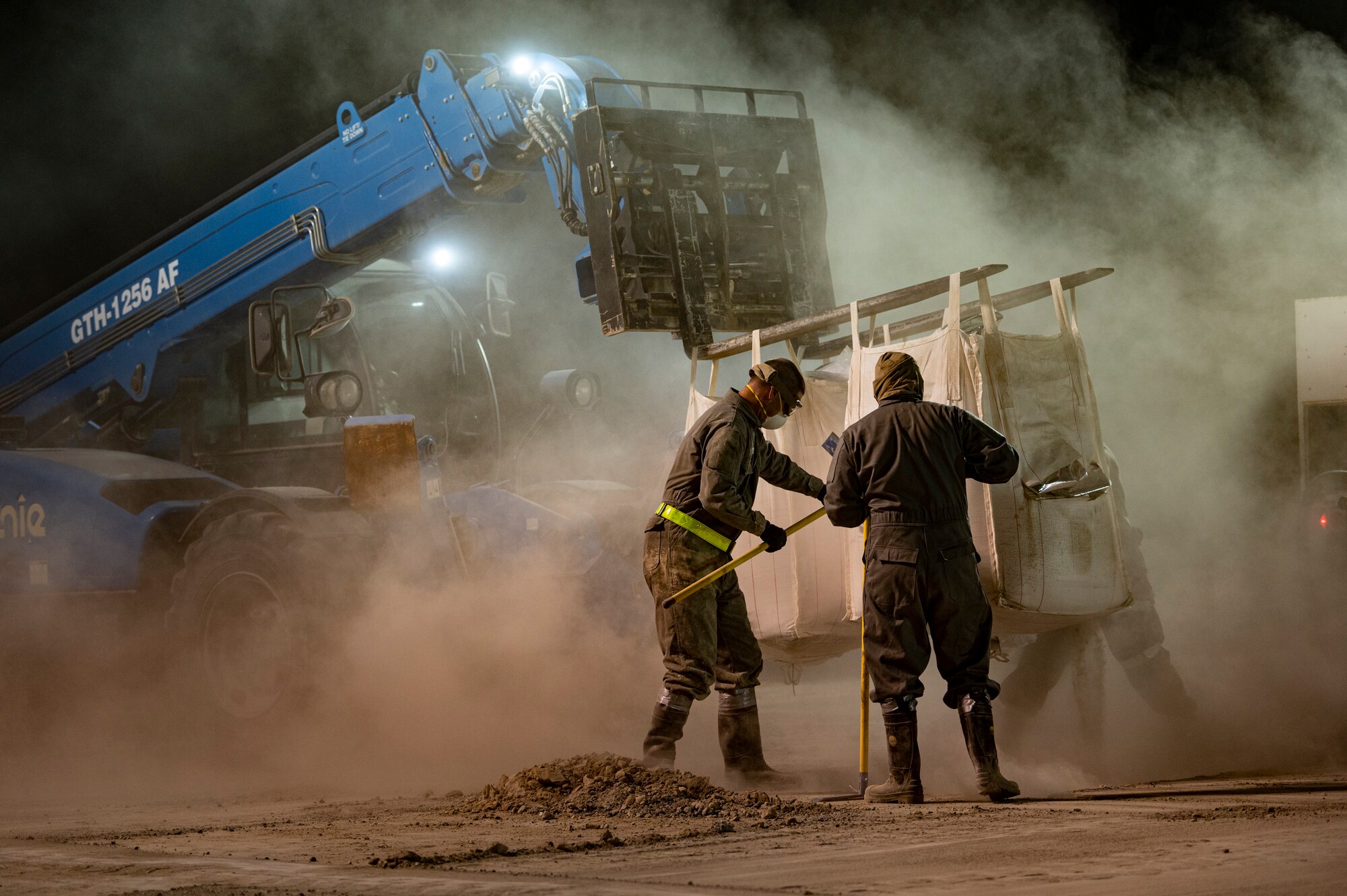 Airmen wearing protective gear fill a hole with dry concrete powder.
