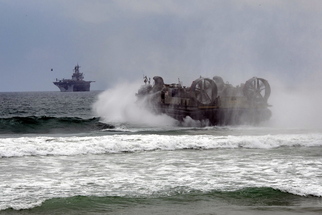 A landing craft leaves the beach while a large military ship floats further out in the ocean.