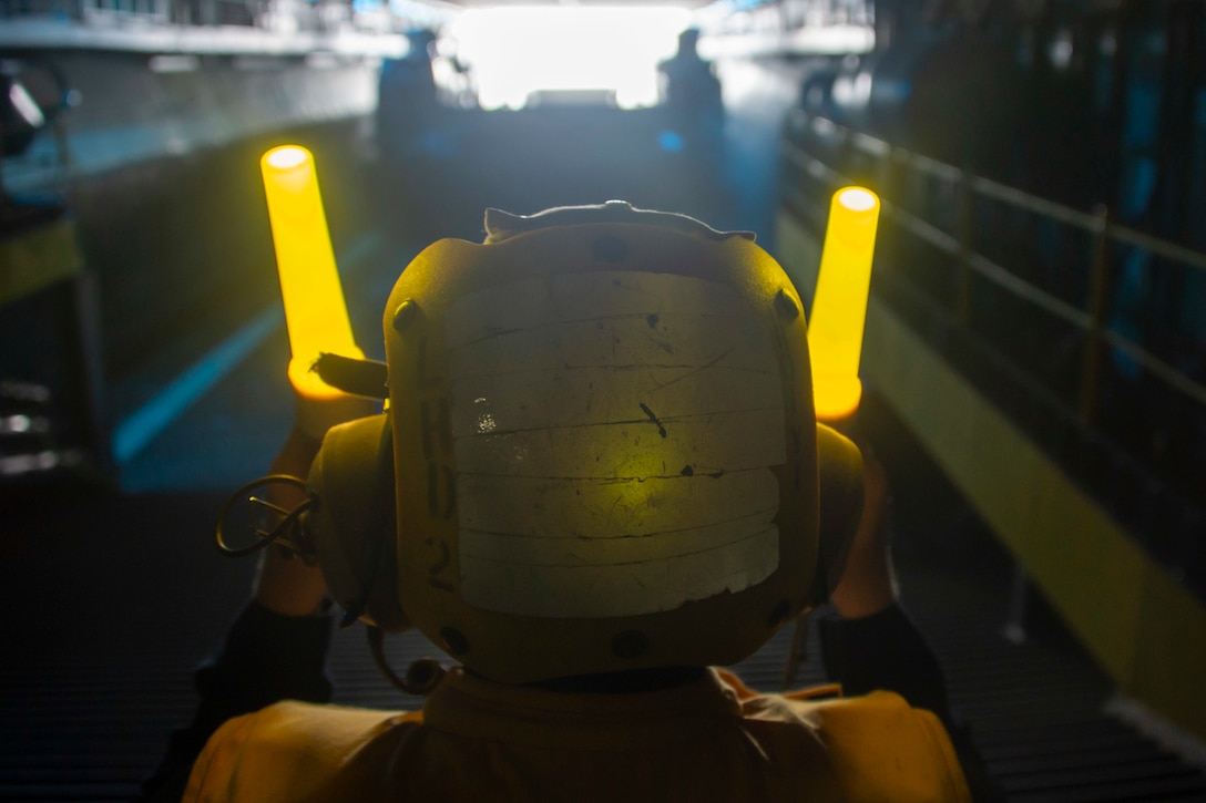 A sailor uses yellow lights to guide a craft into the well deck of a ship.