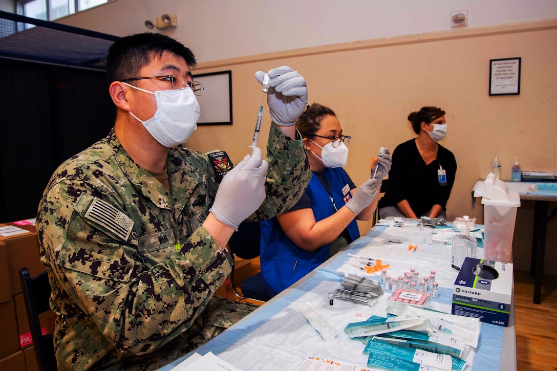 A sailor and registered nurse wearing face masks and gloves hold syringes while preparing them for vaccines at a table while another woman wearing a face mask sits at the end of the table.