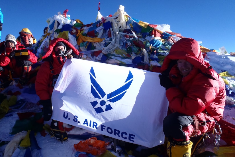 Two bundled-up men hold an Air Force flag atop a peak covered in flags.