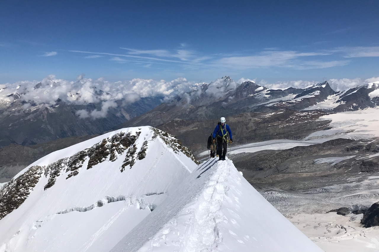 Two men hike along the ridge of a mountain's peak as other mountains loom in the background.